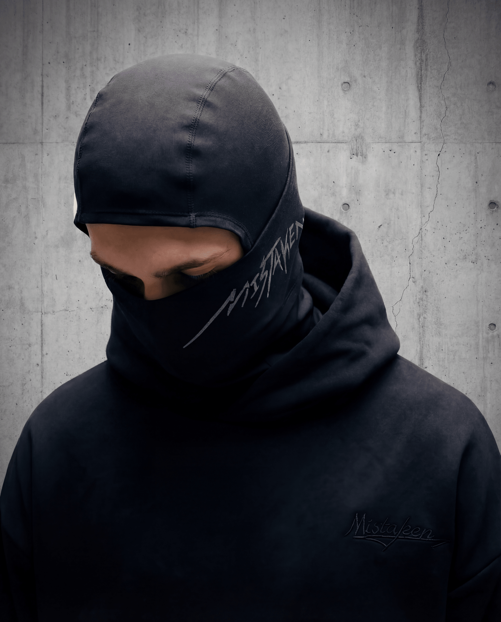 Person wearing a black hooded jacket with a brand logo against a concrete wall.