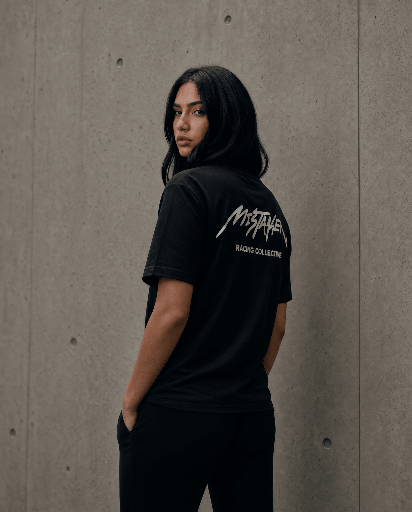 Person wearing a black t-shirt with a logo on the back against a concrete wall.