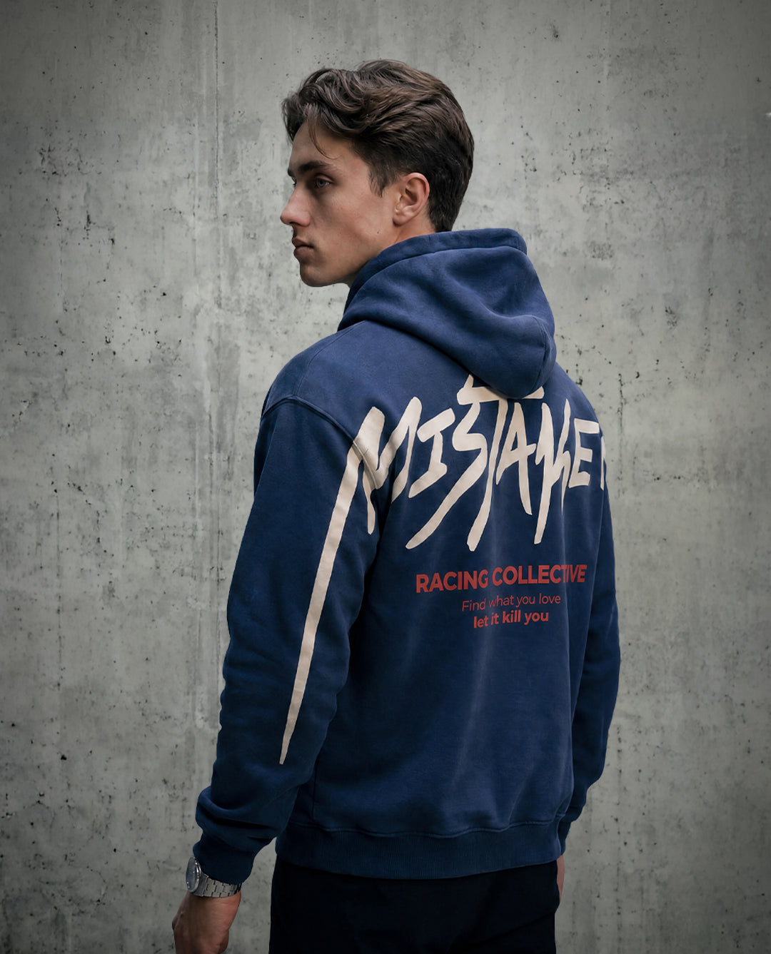 Man wearing a blue hoodie with 'Mistaken' branding against a gray concrete wall.
