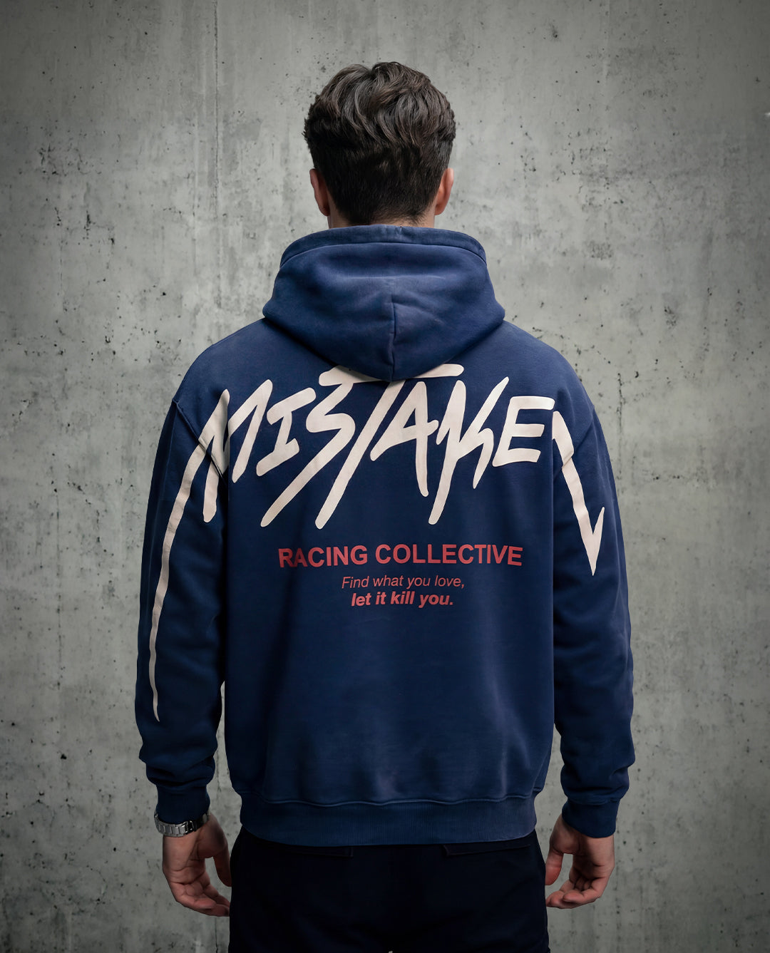 Person wearing a navy hoodie with 'Mistake Racing Collective' text on the back against a gray concrete wall.